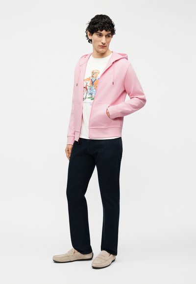 Άντρας που φοράει ανοιχτό ροζ φούτερ με φερμουάρ, άσπρο γραφικό μπλουζάκι με αρκούδα, σκούρο παντελόνι και μπεζ μοκασίνια, στέκεται με το ένα χέρι στην τσέπη.