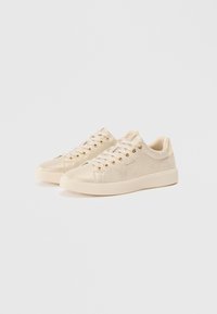 Lyse beige lavtop-sneakers med snøring og gullstifter på hvit bakgrunn, side om side vendt mot høyre.