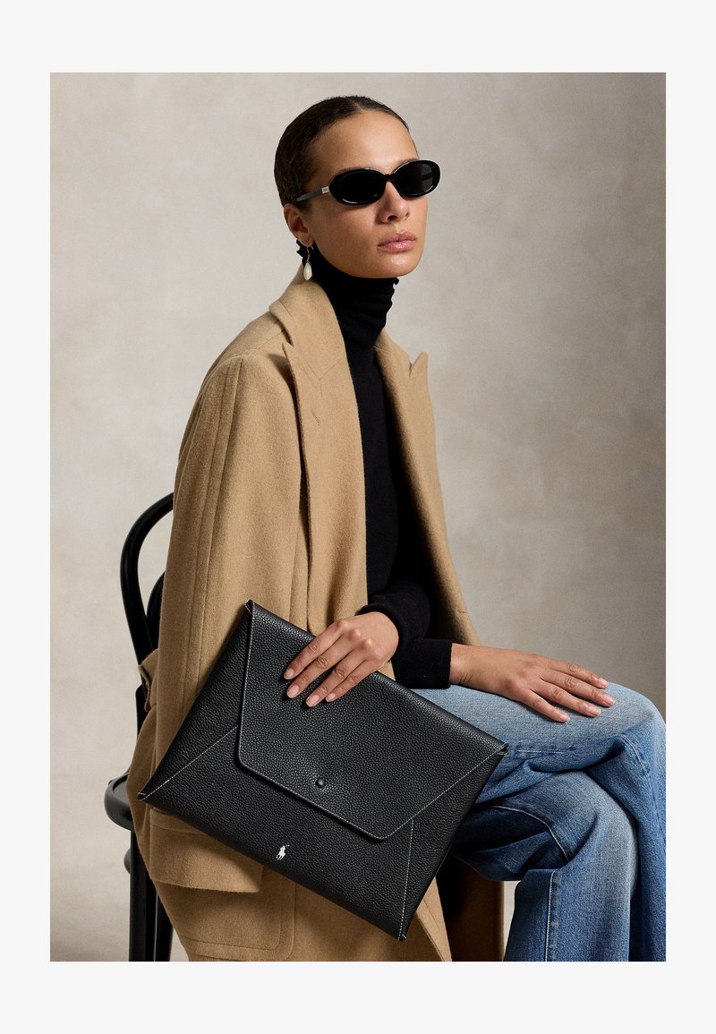 Frau mit schwarzen Sonnenbrillen, beigem Mantel, schwarzem Rollkragenpullover und Jeans, die eine große schwarze Lederumschlag-Clutch hält, während sie auf einem Stuhl sitzt.