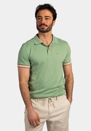 Man draagt een lichtgroen polohemd met korte mouwen en gestreepte manchetten en beige broek met trekkoord, staat met één hand in de zak.