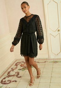 Rochie din dantelă neagră și material transparent cu puncte, cu mâneci lungi, talie strânsă și decolteu în formă de V, asortată cu sandale cu toc înalt și curele aurii.