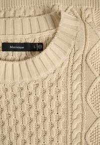 Beige kötött pulóver, texturált dizájnnal, fonott és kábelmintákkal. Bordás nyak és a címke látható a gallér belső részén.