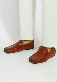Brown leather loafers with a smooth finish, low profile, and stitched details on the upper, paired with light-colored trousers.
