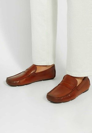 Brown leather loafers with a smooth finish, low profile, and stitched details on the upper, paired with light-colored trousers.