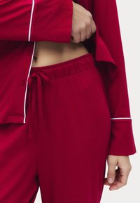 Rød bomulds pyjamassæt med en knap-op top med hvid piping og elastisk taljebukser med snor. Glat stoftekstur.