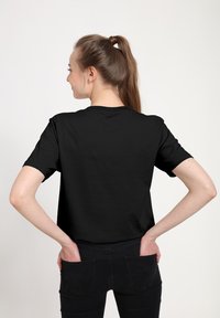 Ung kvinde med hestehale iført en ensfarvet sort t-shirt og sorte bukser, hænderne i baglommerne, vender væk mod en ensfarvet lys baggrund.