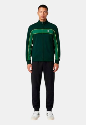 Uomo che indossa una giacca verde scuro con zip e strisce gialle, pantaloni neri e scarpe da ginnastica bianche, in piedi dritto su sfondo semplice.