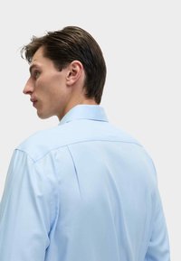 Jasnoniebieska koszula na guziki, gładki materiał, wyraźny kołnierzyk, detal w tylnej części, luźny krój, widoczne szwy na ramionach.