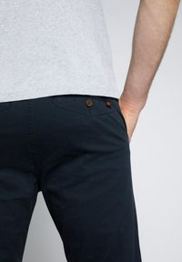 Pantaloni di cotone blu navy con un singolo bottone marrone e cuciture arancioni sulla tasca posteriore, abbinati a una maglietta grigia chiara.