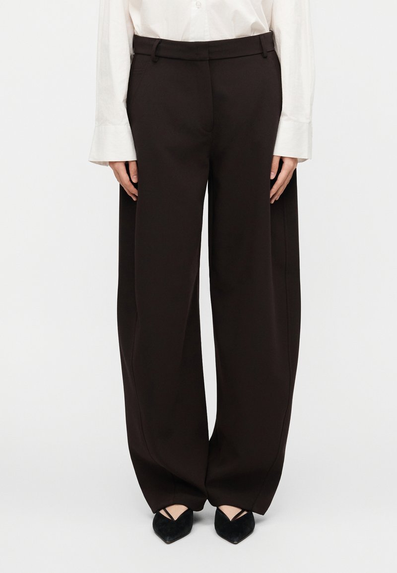 Μαύρο παντελόνι με φαρδύ πόδι από ελαφρύ ύφασμα, με λεία υφή, πιέτες μπροστά και προσαρμοσμένη ζώνη μέσης, συνδυασμένο με μαύρα μυτερά παπούτσια.