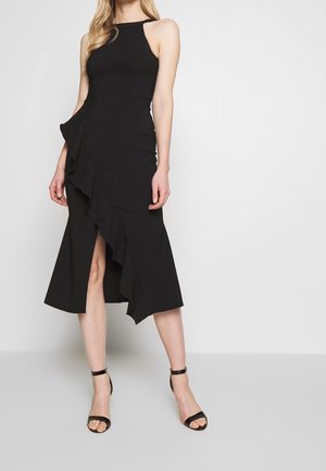 Femme portant une robe midi noire sans manches avec un détail volant asymétrique et des talons hauts noirs à bride à la cheville, debout devant un fond uni.