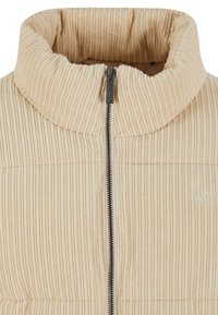 Béžová manšestrová bunda s vysokým límcem a předním zipem, s vertikální žebrovanou texturou a jemným vyšívaným logem.