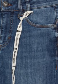 Džinsi no denima vidējā zilā krāsā ar teksturētu virsmu, ar apelsīnu šuvēm, metāla pogu un balto kokvilnas etalonu ar uzrakstu "Organiskā kokvilna".