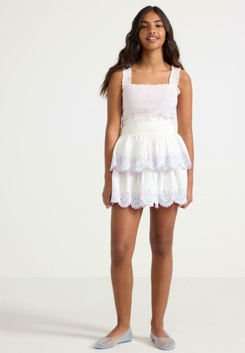 Abito bianco a strati con orlo scalloppato ricamato in rosa e blu, dotato di corpo smock e spalline regolabili. Abbinato a scarpe in rete.