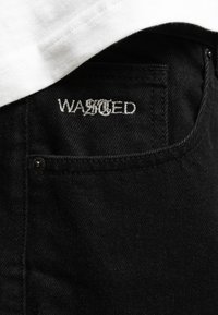 Jeans in denim nero con un primo piano di una tasca con il logo ricamato "WASTED" in filo argento. Il tessuto ha una texture liscia.