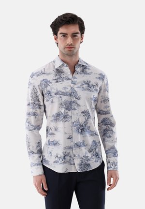 Mann mit dunklem Haar trägt ein weißes Hemd mit Knöpfen und blauem detaillierten Natur- und Tierdruck, kombiniert mit schwarzen Hosen.