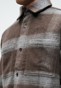 Κασκόλ από παλτό μαλλιού σε καφέ και γκρι καρό σχέδιο με γιακά, πέντε κουμπιά και δύο μπροστινές τσέπες. Υφασμάτινη επιφάνεια με υφή, ζεστή και απαλή στην αφή.