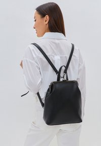 Rucsac din piele neagră, cu o formă structurată, având curele reglabile și o maner prin partea de sus. Textură netedă, cu o închidere metalică cu fermoar.