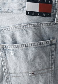 Lyseblå denimjeans med en synlig lomme som har litt flising, samt en merkelapp med teksten "Tommy Jeans" i rødt, hvitt og blått.