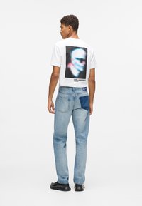 Bijela majica s crnim apstraktnim grafičkim uzorkom na leđima, uparen s svijetloplavim trapericama koje imaju tamnu mrlju. Uključena crna obuća.