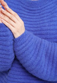 Blauer, gerippter Strickpullover mit langen Ärmeln, der eine weiche, flauschige Textur aufweist. Nahaufnahme von Händen, die auf dem Stoff ruhen.
