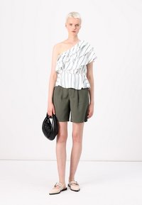 Top alb cu dungi verzi, cu un umăr descoperit și volănașe, combinat cu pantaloni scurți oliv. Modelul ține o geantă de umăr neagră și poartă pantofi deschiși la culoare.