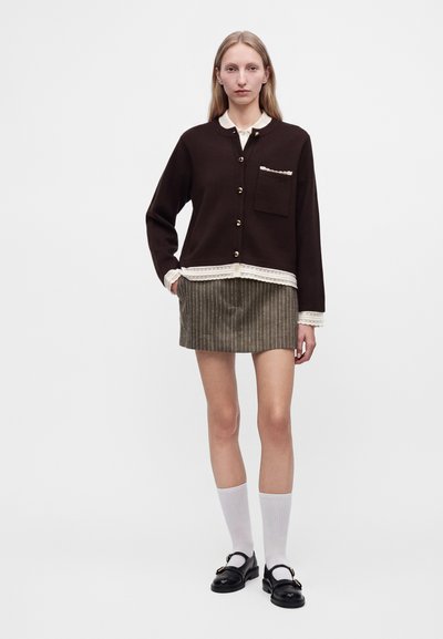 Femme portant un cardigan marron foncé boutonné avec un liseré blanc, une mini-jupe à fines rayures, des chaussettes blanches et des chaussures Mary Jane noires, debout devant un fond blanc.