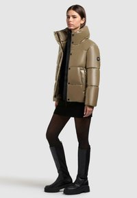 Giubbotto puffer khaki con colletto alto, finitura lucida, dettagli neri e bottoni a pressione, abbinato a un vestito maglione nero e stivali al ginocchio.
