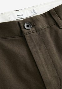 Pantalons droit en brun avec une fermeture à bouton, présentant une surface texturée et des coutures surpiquées. L'étiquette indique le nom de la marque et la taille.