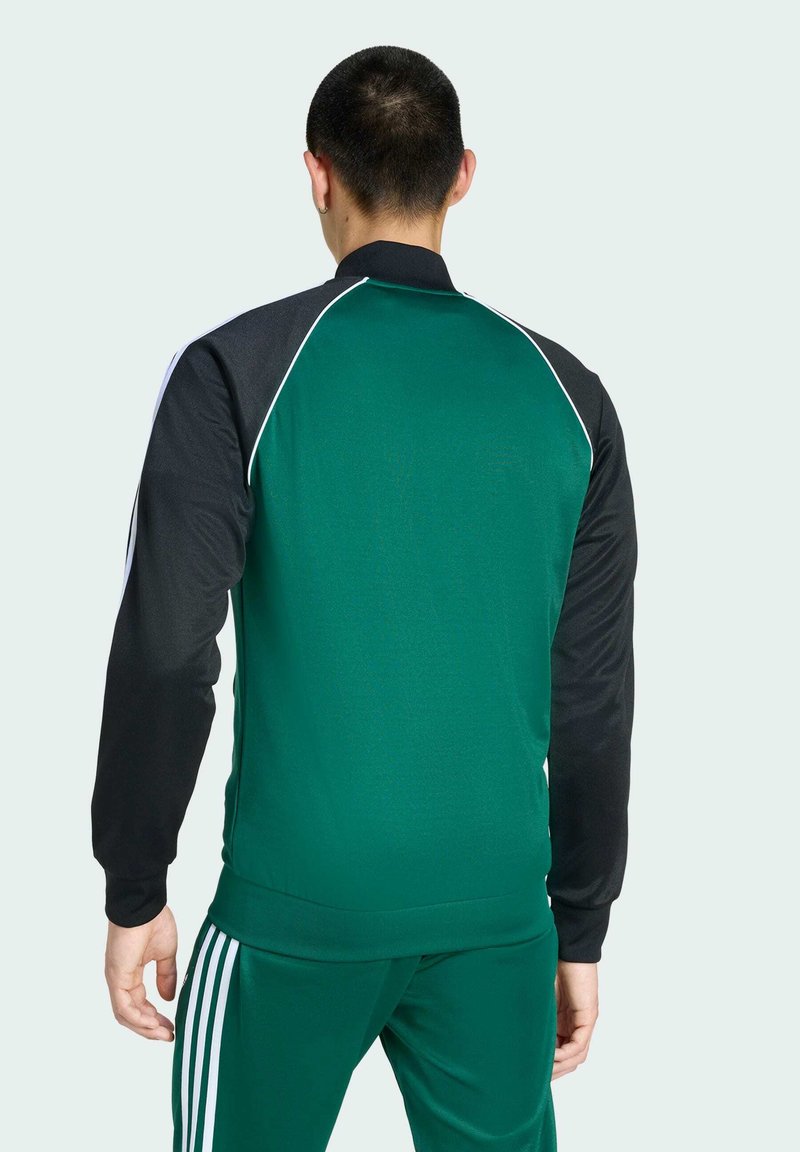 Grön IDrottsjacka med svarta ärmar och vit piping. Slät tyg, figursydd design, ribbad krage och ärmmuddar, har logotyp på baksidan.