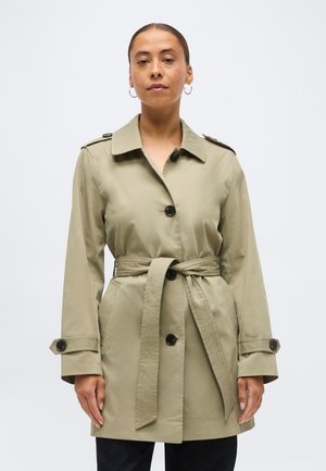 Frau trägt einen beigen Trenchcoat mit Gürtel und schwarzen Knöpfen sowie Creolen, steht vor einem schlichten hellen Hintergrund.