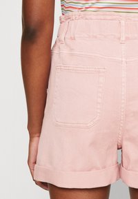 Vaaleanpunaiset farkkushortsit kääntösaumalla, jossa on takatasku. Materiaali on pehmeä ja siinä on näkyvällä saumanomistustyylillä.