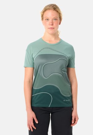 Frau mit lockigem blondem Haar, die ein grünes Oberteil mit abstrakten Konturlinien und schwarze Hosen trägt, steht vor einem schlichten Hintergrund.