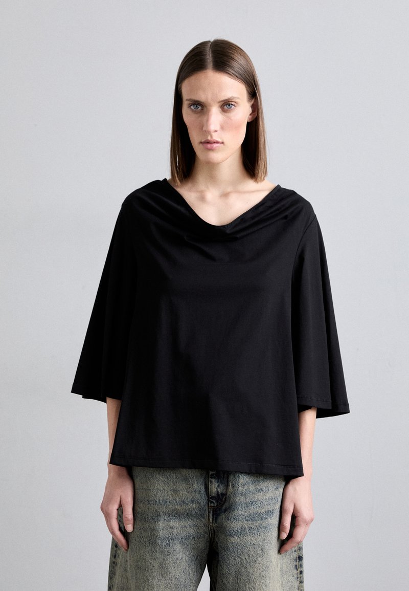 Black short-sleeved top with a wide, draped neckline. Made of soft fabric, featuring a relaxed fit. Paired with light blue, loose-fitting jeans.