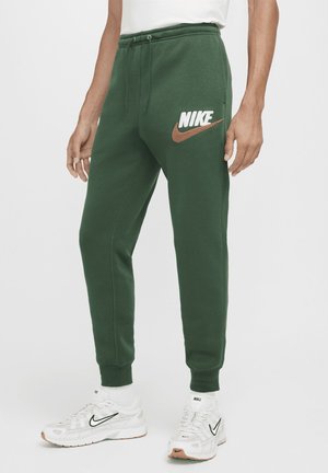 Personne portant un pantalon de survêtement Nike vert avec cordon et des baskets blanches Nike avec un swoosh noir sur un fond uni.
