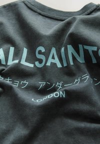 T-paita tummanharmaata puuvillaa, jossa on ryppyinen rakenne. Siinä on vaaleansinaiset, rohkealla fontilla kirjoitetut tekstit "ALLSAINTS" ja "LONDON", sekä lisäsymboleita kontrastoivilla tyyleillä.