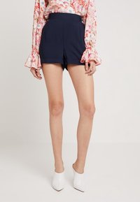 Shorts taille haute bleu marine avec plis avant, design structuré et texture lisse, assortis à un chemisier à manches longues à motifs floraux et des talons blancs.