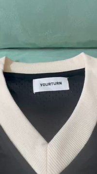 V-veida džemperis ar melnu ķermeni un krēmkrāsas ribotu apkakli. Iekšpusē ir "YOURTURN" birka. Materiāls izskatās viegls un elpojošs.