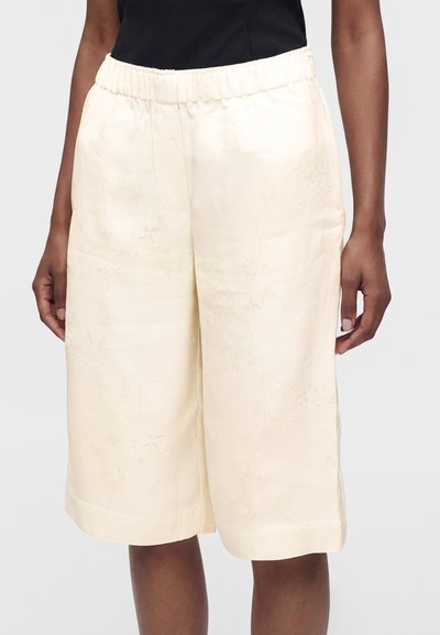 Femme portant un short crème au genou avec un motif floral subtil et une taille élastique, sur un fond gris uni.