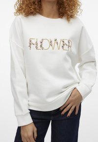 Vit sweatshirt med texten "FLOWER" i broderat guld och rosa blommönster. Har neddragna axlar och ribbade mudd. Mjukt tyg.