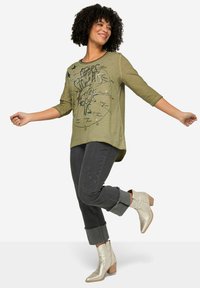 Olivenfarbenes Langarmshirt mit grafischem Druck, kombiniert mit dunklen Jeans mit dekorativen Akzenten und silbernen Ankle Boots mit einem gestapelten Absatz.