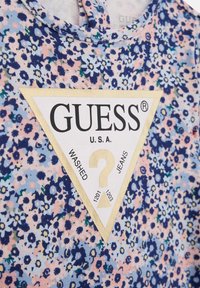Camicia a fantasia floreale in tonalità di blu e rosa, con un'etichetta triangolare bianca con testo nero e dorato per "GUESS", completa di un dettaglio a forma di punto interrogativo.