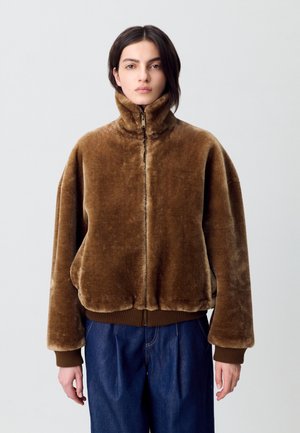 Chaqueta de pelaje sintético marrón con cuello alto, cierre de cremallera y puños acanalados. Presenta un corte holgado y una textura suave; llevada sobre pantalones de mezclilla azul.