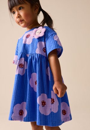 Junges Mädchen mit dunklem Haar in einem blauen Kleid mit großen rosa Blumen und einem lächelnden Blumenaufnäher auf der Brust, vor einem schlichten Hintergrund stehend.