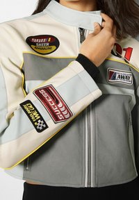 Veste de moto en cuir de couleur gris clair et crème avec des accents jaunes. Comprend plusieurs patches brodés et une fermeture éclair à l'avant.