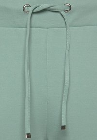 Helle grüne Sweatpants im Nahaufnahme, mit einem Kordelzug und metallischen Aglets sowie einer glatten, strukturierten Stoffoberfläche.
