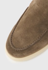 Καφέ μοκασίνι από σουέτ με λεπτομερή ραφή και ελαφριά μπεζ σόλα από καουτσούκ σε λευκό φόντο.