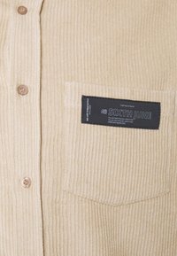 Beżowa koszula ze sztruksu o teksturowanej powierzchni, z sześcioma guzikami i naszywaną kieszenią z czarną metką.