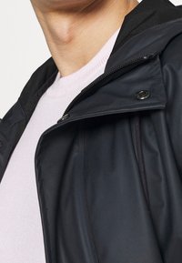 Svart jacka med huva, med dragkedja och tryckknappar, bärs över en ljusrosa tröja. Materialet verkar slätt och lätt.