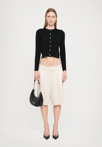 Black cropped cardigan with gold buttons, cream pleated shorts, black handbag with chain handle, and pointed toe black heels.